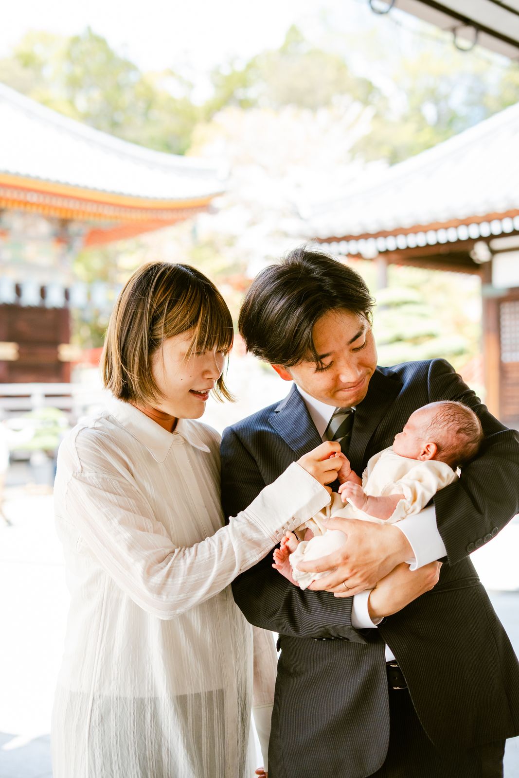 お宮参りで神社を背に新生児を抱くご夫婦の写真